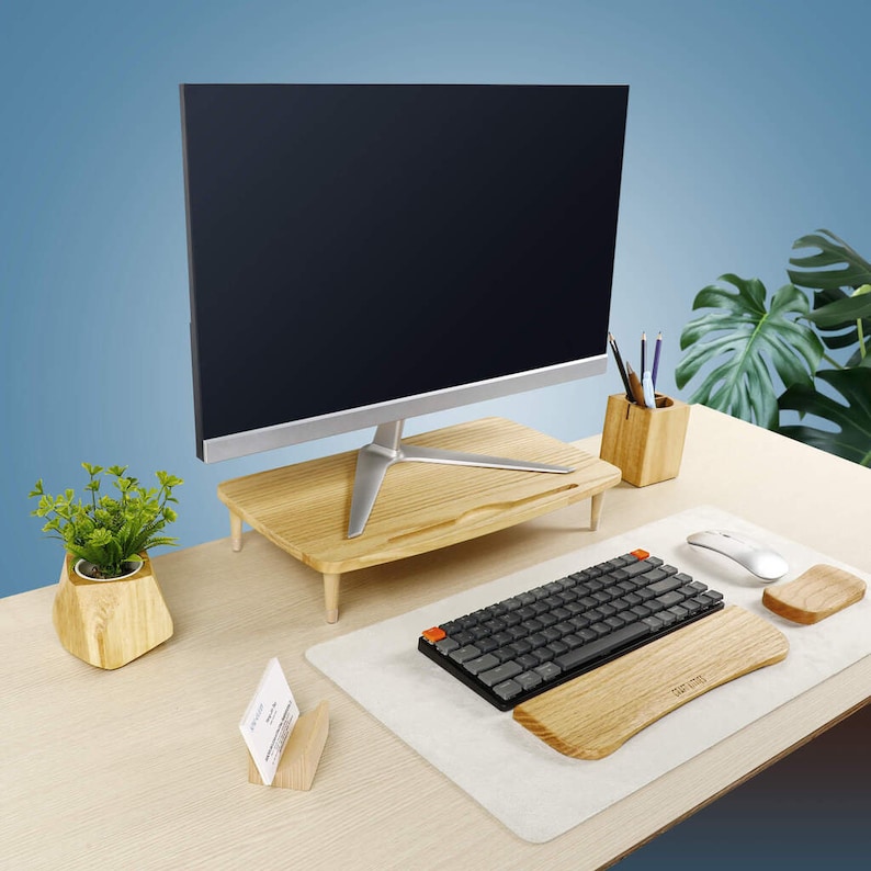 Pu&ograve; includere: Un supporto per monitor in legno con un monitor per computer nero in cima. Un tappetino da scrivania bianco con una tastiera nera e un poggiapolsi in legno. Un portapenne in legno con matite e una piccola pianta in vaso su una scrivania in legno.