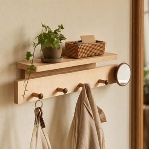 Puede incluir: Un estante de pared de madera de tono claro con una repisa superior, cuatro ganchos para colgar objetos y un pequeño espejo circular en el lado derecho. Una cesta rectangular tejida y una planta verde se encuentran sobre el estante.