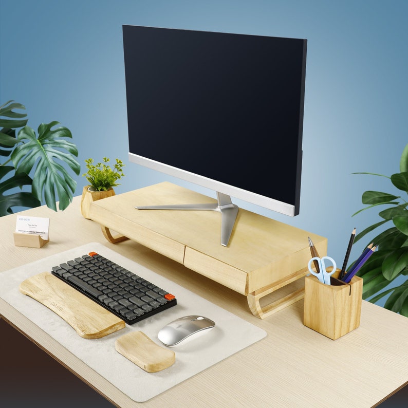 May include: A wooden monitor stand with a drawer, a black keyboard, a wooden wrist rest, a white mouse, and a wooden pencil holder with pencils and a pair of scissors. The stand is on a light wood desk.