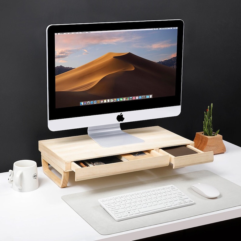 May include: A wooden monitor stand with two drawers, a white keyboard, a white mouse, and a white iMac computer with a desert landscape on the screen. The stand is on a white desk.