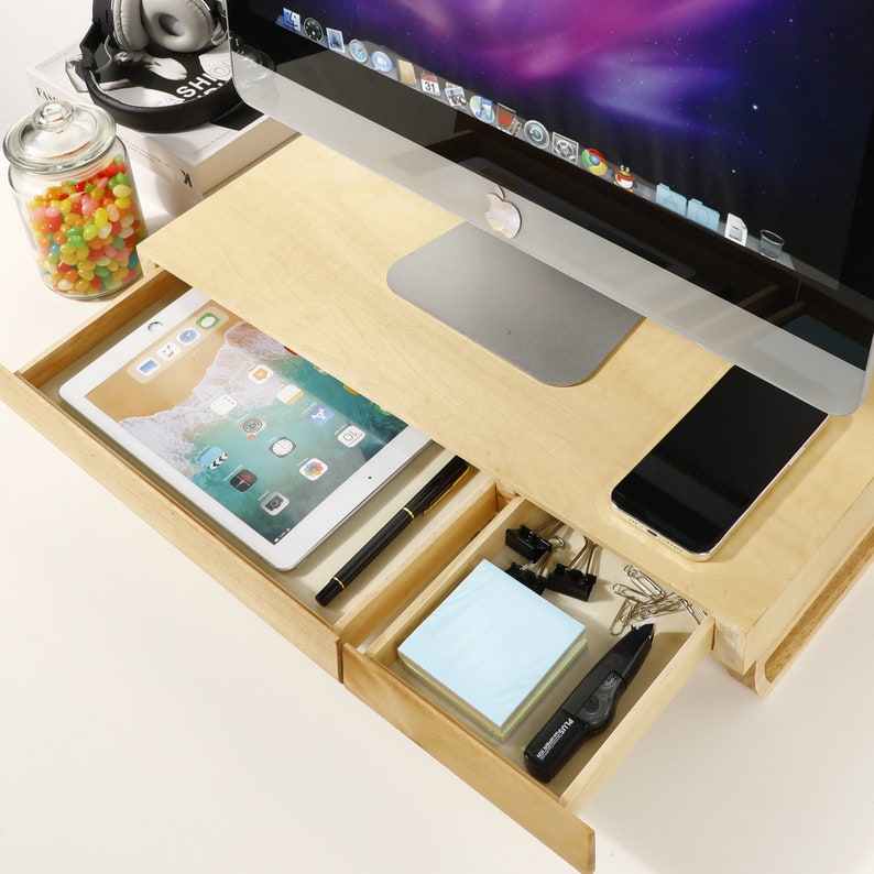 May include: A wooden monitor stand with a drawer that opens to reveal a tablet, a pen, and a stack of sticky notes. There is a smartphone and a set of paper clips in the other drawer.