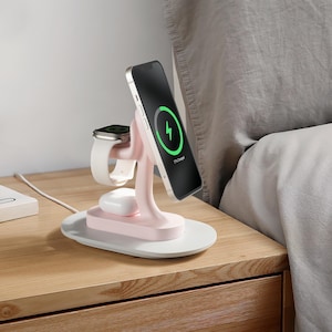 May include: A pink wireless charging stand with a white base. The stand has three charging pads: one for a phone, one for a smartwatch, and one for wireless earbuds. The phone is charging and shows a green charging indicator on the screen. The stand is on a wooden nightstand.