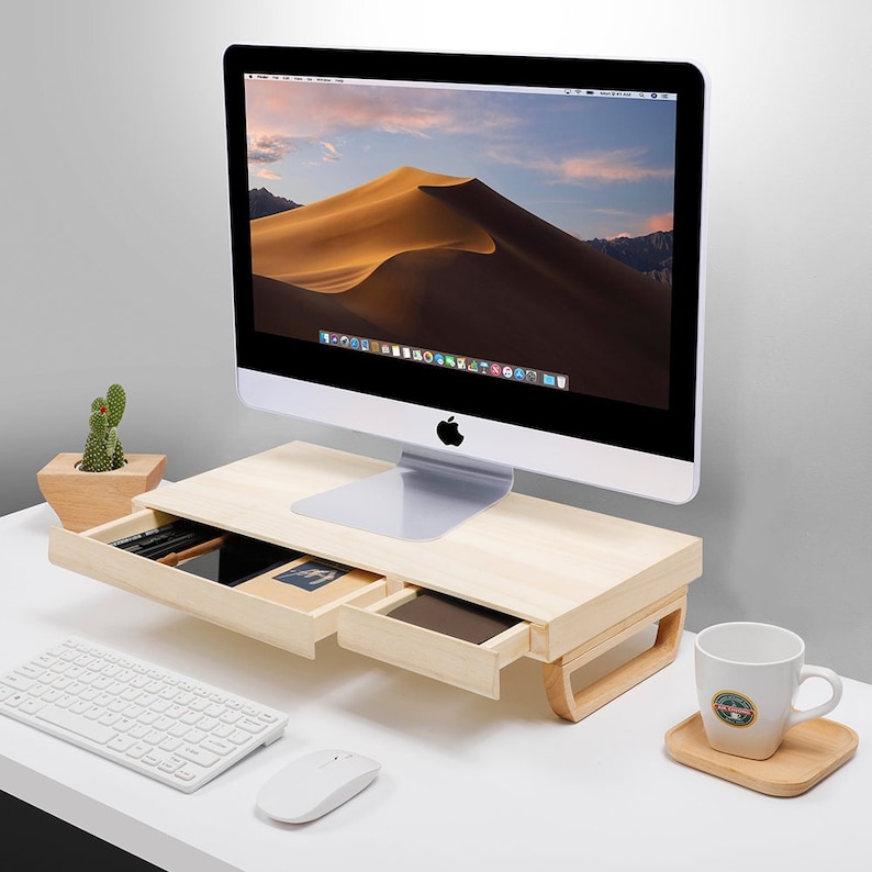 May include: A wooden monitor stand with two drawers for storing office supplies. The stand is made of light wood and has a modern design. A computer monitor, keyboard, mouse, and a coffee cup are on the desk.