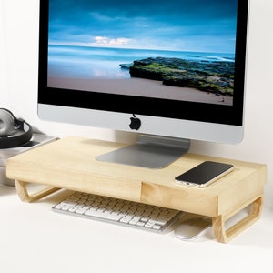 May include: A light wood monitor stand with a drawer for storing items like a keyboard, mouse, or phone. The stand is raised to provide ergonomic support for a computer monitor.