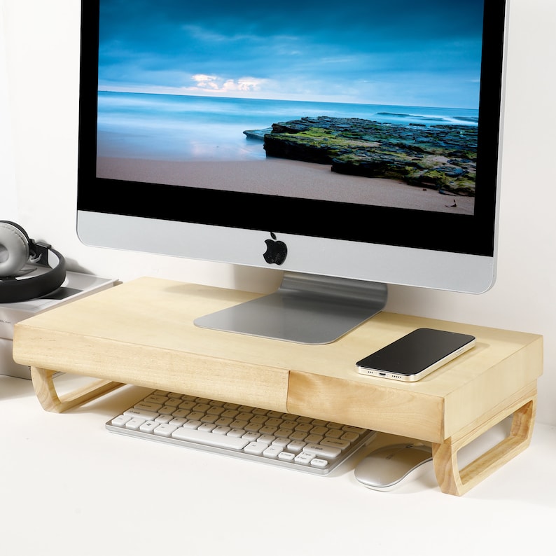 May include: A light wood monitor stand with a drawer for storing items like a keyboard, mouse, or phone. The stand is raised to provide ergonomic support for a computer monitor.