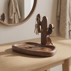 Peut inclure: Un plateau organisateur en bois brun foncé doté d'un support en forme de cactus avec de petits crochets en métal doré. Le plateau contient deux petites pièces dorées, tandis que le support cactus présente un trousseau de clés et des lunettes.