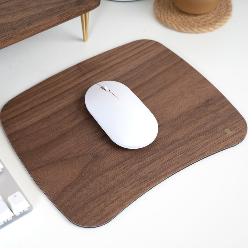 May include: A brown wood mouse pad with a white wireless mouse on top. The mouse pad has a curved edge and the text "CRAFTSMITH" in gold lettering in the bottom right corner.