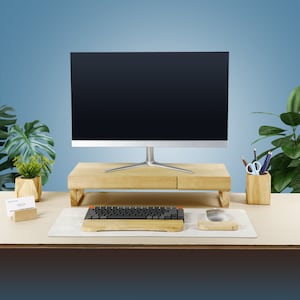 May include: A wooden monitor stand with a black keyboard and a white mouse on a light brown desk. The monitor stand has a drawer and is raised on two legs. There are two potted plants and a business card holder on the desk.