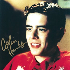 May include: A close-up portrait of a young man with short brown hair wearing a red shirt with white embroidery. The man is smiling and looking at the camera. The image is signed "Colin Hanks".