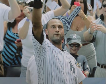 May include: A baseball player in a white and blue striped jersey with the number 6 on the sleeve raises his right arm in the air, pointing his index finger towards the sky. The player is wearing a black belt and white pants. The player is surrounded by fans in the stands.