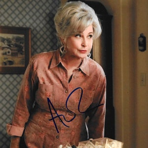 May include: A woman with short, gray hair wearing a pink floral button-down shirt and black pants. She is standing in a room with a clear plastic storage container filled with various items. The woman has signed her name in blue ink on the shirt.
