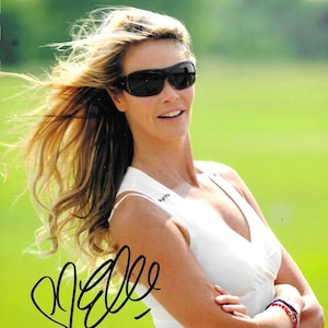 Può includere: Una donna con lunghi capelli biondi che indossa occhiali da sole e una camicia bianca senza maniche con un logo nero. Sorride e tiene le braccia incrociate. Lo sfondo è un campo erboso verde. L'immagine è firmata "Elle".