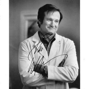 May include: A black and white photograph featuring a person with short dark hair wearing a collared shirt under a white lab coat. The individual has arms crossed and is looking slightly to the side. A black ink signature is across the chest area.