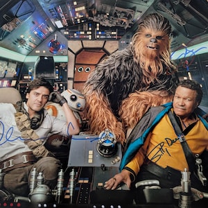 May include: A Star Wars film still featuring Chewbacca, Oscar Isaac, and Billy Dee Williams in a spaceship cockpit. The image is signed by the actors. The cockpit is filled with buttons, screens, and other controls.