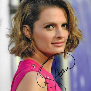 May include: A portrait of a woman with short, wavy brown hair. She is wearing a bright pink dress with decorative clasps on the shoulder. A black signature is visible on the image.
