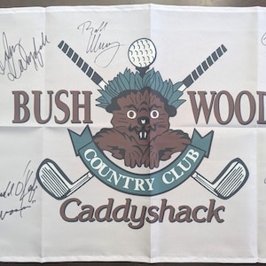 May include: A white golf flag featuring the Bushwood Country Club logo with a brown gopher, crossed golf clubs, and a golf ball. The text reads Bushwood Country Club Caddyshack. Several black ink signatures are visible across the flag surface.