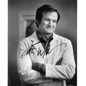 May include: A black and white photograph featuring a person with short dark hair wearing a collared shirt under a white lab coat with arms crossed. A bold black ink signature is written across the center of the image over the lab coat.
