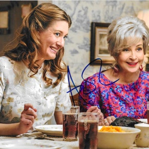 May include: Two women seated at a table, smiling. One wears a floral dress, the other a red and blue floral top. Glasses of a dark beverage and plates of food are on the table. An autograph is visible on the image.