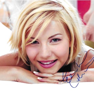 May include: A close-up portrait of a woman with short blonde hair. She is smiling and looking at the camera. Her signature is visible in the bottom right corner of the image.