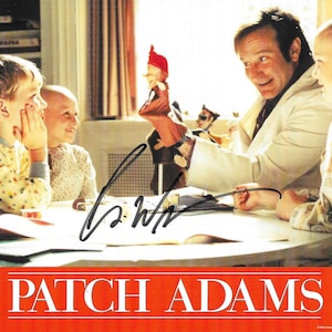 Könnte beinhalten: Ein Werbefoto für den Film Patch Adams, das eine Person im weißen Kittel zeigt, die mit einer roten Handpuppe Kinder an einem Tisch unterhält. Das Bild enthält ein Autogramm in schwarzer Tinte und den Text PATCH ADAMS am unteren Rand.