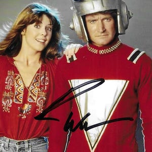 Könnte beinhalten: Ein signiertes Foto mit einer Person in einem roten Langarmshirt mit weißem, dreieckigem Brustbereich, goldenem Kragen und silbernem Helm, neben einer Person in einer roten Bluse mit gemustertem Ausschnitt und einer blauen Jeans.
