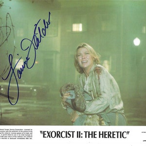 May include: A movie poster for the film "EXORCIST II: THE HERETIC" featuring a woman in a white shirt holding a child. The woman has a distressed look on her face and is surrounded by a green fog. The poster is signed by actress Louise Fletcher.