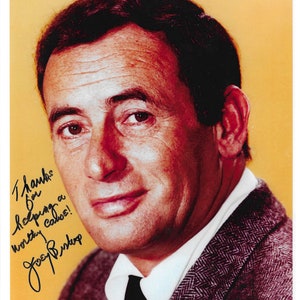 Peut inclure: Une photo en noir et blanc d'un homme aux cheveux foncés portant une veste de costume marron. Il sourit et regarde la caméra. L'homme tient un stylo et signe son nom, "Joey Bishop", sur un morceau de papier. Le texte "Thanks for helping a worthy cause!" est écrit au-dessus de sa signature.