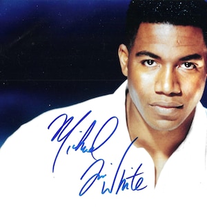 以下が含まれることがあります： 白い襟付きシャツを着た、短い黒髪の男性の白黒写真。男性はカメラを見ており、真剣な表情をしています。写真は青いインクで「Michael Jai White」と署名されています。