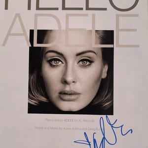 Puede incluir: Portada de partitura de "Hello" de Adele, con un retrato en blanco y negro de la cantante. El título "HELLO ADELE" está impreso en letras grandes. Incluye la firma del artista en tinta azul.