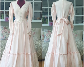 May include: A long-sleeved, light pink dress with a lace trim neckline and a bow at the back. The dress has a tiered skirt and is made of a soft, flowing fabric.