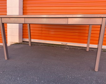 May include: A silver metal desk with two drawers. The desk is in front of an orange garage door.