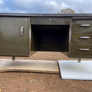 May include: A dark green metal desk with a single drawer and three drawers on the right side. The desk has a simple design with a flat top and a smooth surface.