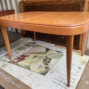 Puede incluir: Una mesa ovalada de madera clara con una superficie lisa y pulida y cuatro patas delgadas. La mesa está sobre una superficie con estampado marrón y blanco.