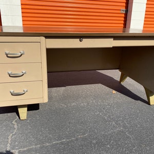 May include: A vintage metal desk with a light beige finish and four drawers. The desk has a single drawer in the center and three drawers on the left side. The desk has gold-colored legs.