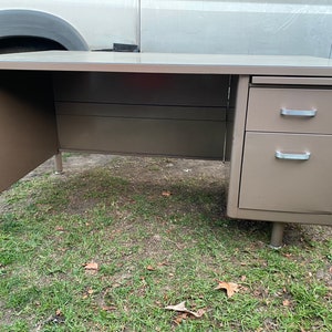 May include: A vintage metal desk in a neutral taupe color. The desk features a large work surface, a two-drawer cabinet, and four cylindrical legs. The drawers have silver-colored handles. The desk is designed for office or home use.