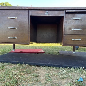 May include: A vintage wooden desk with a dark brown finish. It features a central open space for a chair, flanked by cabinets and drawers with silver-toned handles. The desk is supported by four legs with silver caps.
