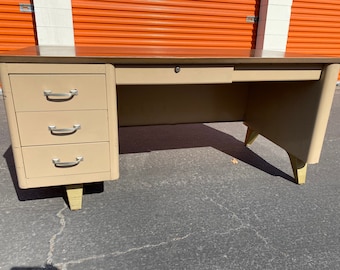 May include: A vintage metal desk with a light beige finish and four drawers. The desk has a single drawer in the center and three drawers on the left side. The desk has gold-colored legs.