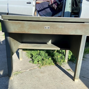 May include: An industrial-style metal desk in a muted green color. The desk has a rectangular top, a single drawer, and a lower shelf. The legs are sturdy and square, with a small emblem on the front. The desk is designed for functionality and durability.