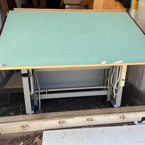 May include: A large drafting table with a light green surface and a wooden frame. The table has a metal base and an attached power strip. The table is inside a shipping container.