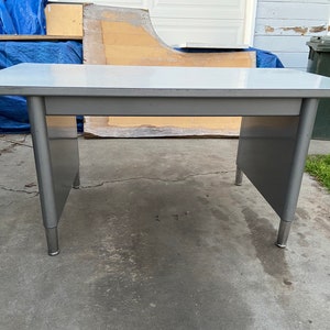 May include: A gray metal desk with a white top and rounded edges. The desk has four legs and is sitting on a concrete surface.