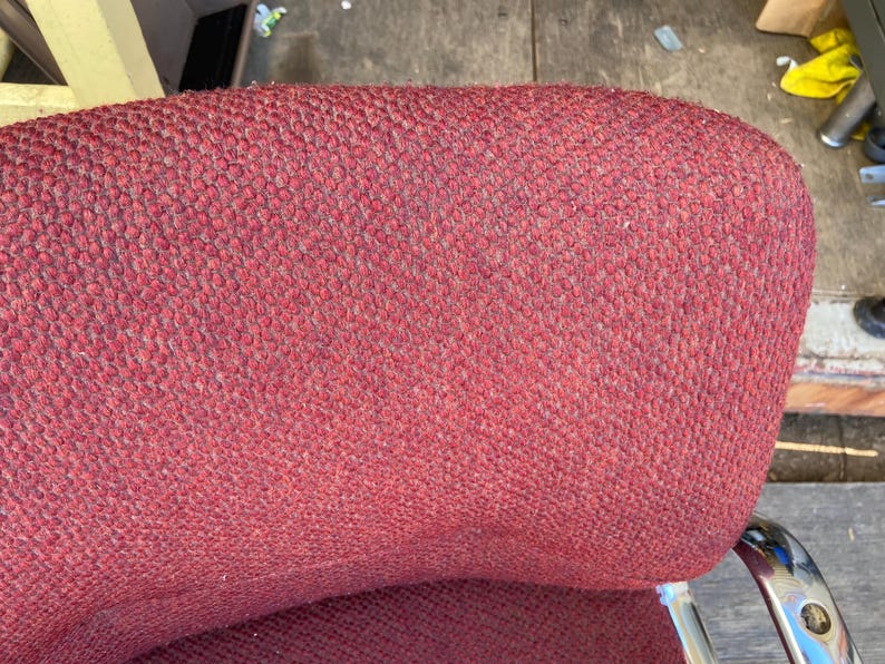 Puede incluir: Primer plano del respaldo de una silla tapizada roja. La tela tiene un aspecto texturizado y tejido. La silla tiene un respaldo curvo y un marco de metal cromado. La silla parece ser vintage.