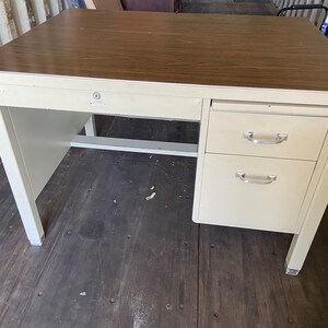 May include: A vintage metal desk with a wood-grain laminate top. The desk has a cream-colored frame, two drawers, and a single drawer. The desk has a keyhole and silver-colored drawer pulls. The desk is rectangular and appears to be sturdy.