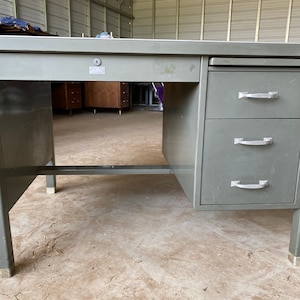 May include: A gray metal desk with three drawers on the right side. The desk has a single drawer on the left side with a keyhole. The desk has a metal frame and legs.