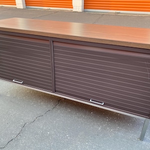 Puede incluir: Un mueble moderno de mediados de siglo con una tapa de madera marrón y un frente de listones marrón oscuro. El mueble tiene dos puertas correderas con tiradores plateados y descansa sobre patas de metal plateado. Una solución de almacenamiento elegante.