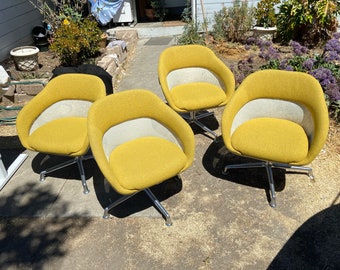 May include: Four mid-century modern swivel chairs. The chairs have a mustard yellow fabric seat and back with a white mesh center. The base is chrome with a five-star design. The chairs are on a concrete surface.