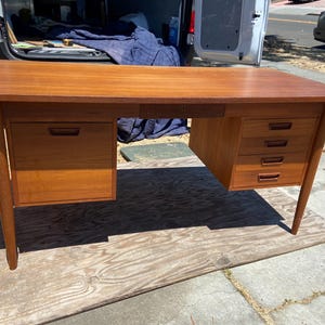 May include: A mid-century modern wooden desk with a warm, reddish-brown finish. The desk features two sets of drawers, one with two drawers and the other with four, and a central drawer. The desk is supported by angled legs.