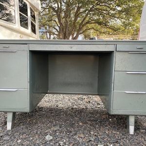 May include: A vintage metal desk in a muted green color. The desk has multiple drawers with silver handles and a keyhole. The desk is supported by silver-capped legs. The desk is designed for office or home use.