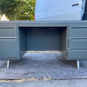 May include: A vintage gray metal desk with two sets of drawers and a central open space. The desk has a rectangular shape, with silver legs and handles. The desk is likely from the mid-century era.