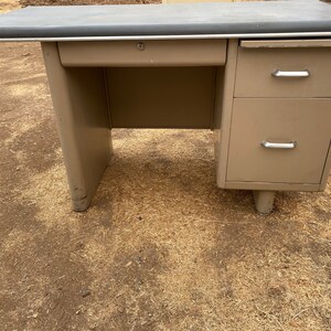 May include: A vintage metal desk with a light brown finish. The desk has a single drawer on the left side and two drawers on the right side. The desk has a dark gray top and silver drawer pulls.