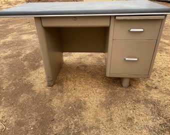 May include: A vintage metal desk with a light brown finish. The desk has a single drawer on the left side and two drawers on the right side. The desk has a dark gray top and silver drawer pulls.